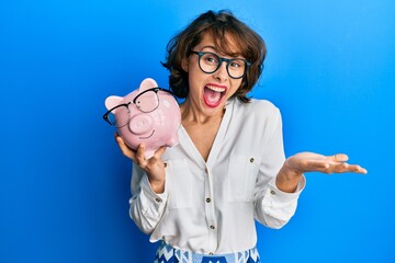 Young brunette woman holding piggy bank with glasses celebrating achievement with happy smile and winner expression with raised hand