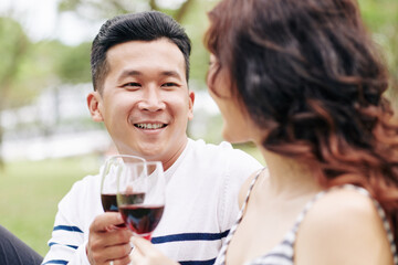 Portrait of smiling handsome young Asina man enjoying picnic with girlfriend and drinking wine