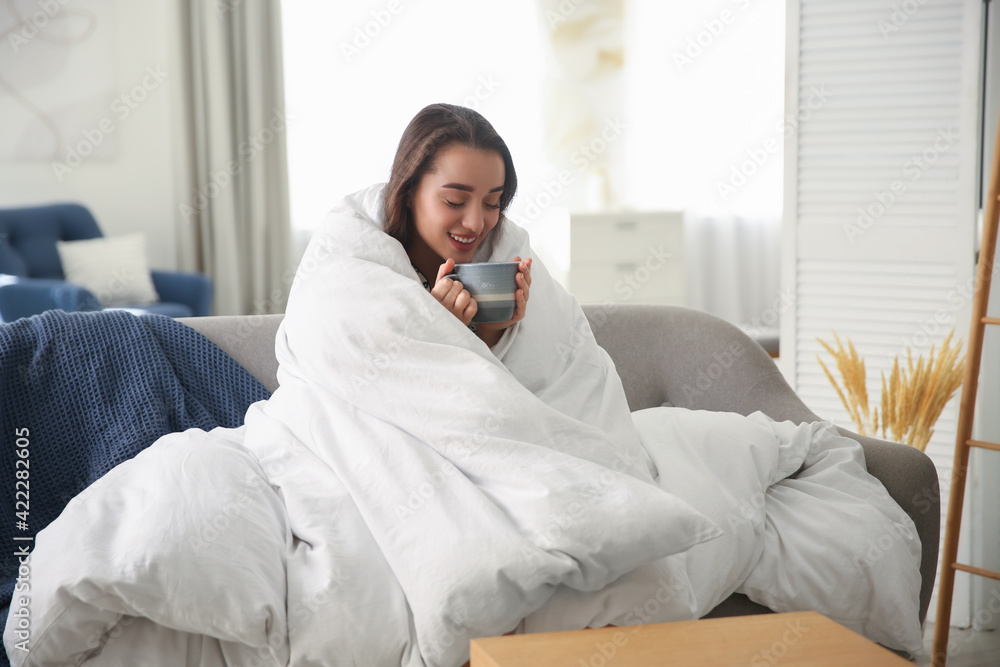 Canvas Prints Beautiful young woman wrapped with soft blanket holding cup of hot drink on sofa at home