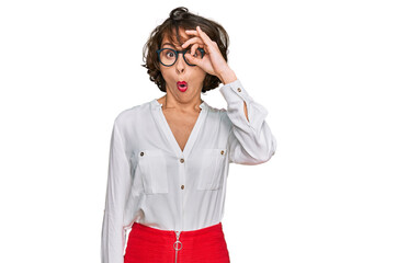Young hispanic woman wearing business style and glasses doing ok gesture shocked with surprised face, eye looking through fingers. unbelieving expression.