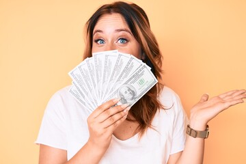 Young beautiful caucasian woman holding dollars celebrating achievement with happy smile and winner expression with raised hand