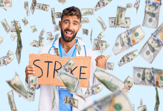 Young hispanic man wearing doctor stethoscope holding strike banner pointing thumb up to the side smiling happy with open mouth
