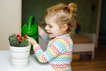 Little toddler girl watering flowers and cactus plants on window at home. Cute child helping, domestic life. Happy healthy kid holding water can, leaning help. Greenery, environment concept.