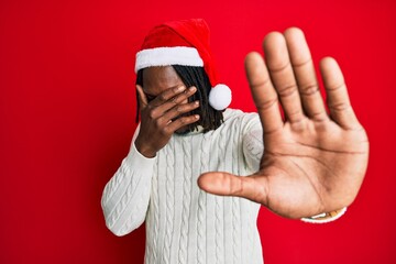 African american man with braids wearing christmas hat covering eyes with hands and doing stop gesture with sad and fear expression. embarrassed and negative concept.