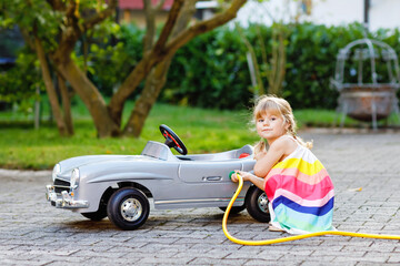 Little toddler girl playing with big vintage toy car and having fun outdoors in summer. Cute child refuel car with water. Girl using garden hose and fill up with gasoline, role game gas station. - obrazy, fototapety, plakaty