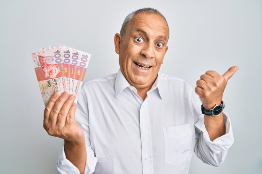 Handsome senior man holding 100 hong kong dollars banknotes pointing thumb up to the side smiling happy with open mouth