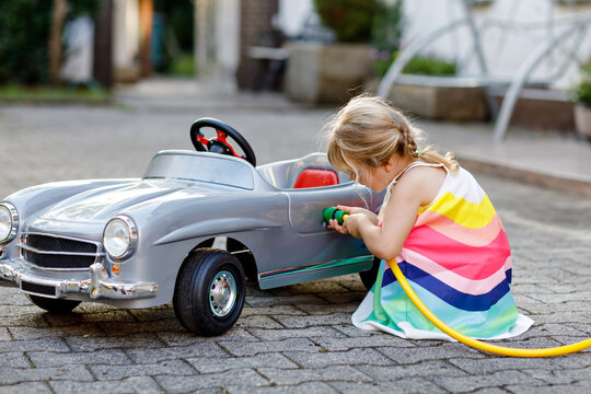 Little toddler girl playing with big vintage toy car and having fun outdoors in summer. Cute child refuel car with water. Girl using garden hose and fill up with gasoline, role game gas station.
