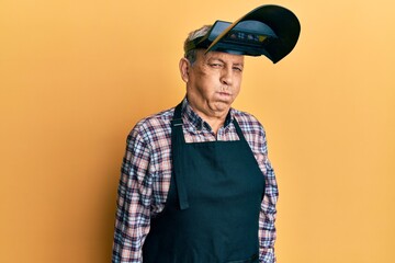 Handsome senior man with grey hair wearing welding protection mask puffing cheeks with funny face. mouth inflated with air, crazy expression.