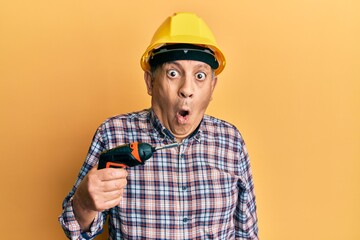 Handsome senior man with grey hair holding screwdriver wearing hardhat scared and amazed with open mouth for surprise, disbelief face