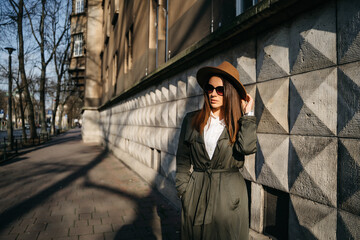 Outdoor fashion portrait of elegant, luxury woman wearing beige hat, sunglasses, trendy white shirt, in a green trench coat, walking in street of European city. Copy, empty space for text