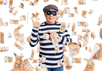 Senior handsome man wearing burglar mask and t-shirt pointing to the back behind with hand and thumbs up, smiling confident