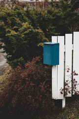 Blue mailbox in the front of the house. White fence and green bushes.