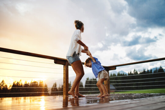 Mother with small daughter playing in rain on patio of wooden cabin, holiday in nature concept.