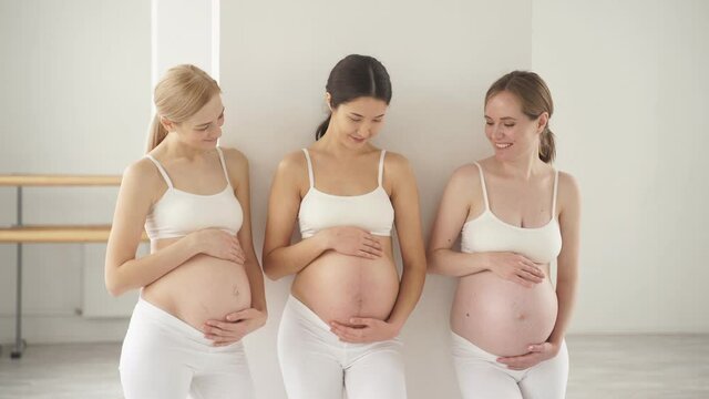 Three young pregnant women standing in studio in white sportive outfit with naked tummy, future moms stand talking, hugging their belly.