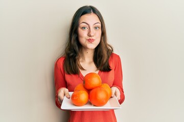 Young brunette woman holding plate with fresh oranges puffing cheeks with funny face. mouth inflated with air, catching air.