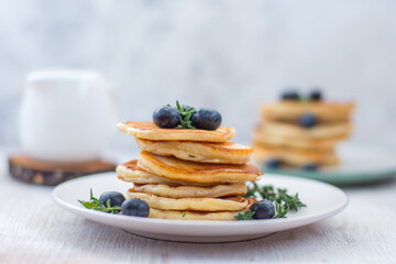 Pancakes laid out in a stack on a plate with blueberries and decorated with herbs on a light background. The background is blurred. Horizontal arrangement