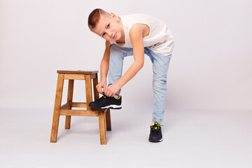 Nine-year-old European boy puts his foot on stool and ties his shoelaces on a white background