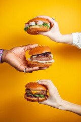 Hands holding burger against yellow background. Fast food, junk food concept. - obrazy, fototapety, plakaty