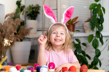 a little blonde girl sits at the table and paints Easter eggs with brushes and paints, smiles, on the head of the ears of the Easter bunny. the concept of a religious holiday and spring