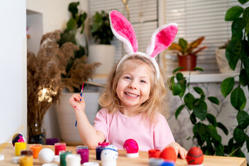 a little blonde girl sits at the table and paints Easter eggs with brushes and paints, smiles, on the head of the ears of the Easter bunny. the concept of a religious holiday and spring