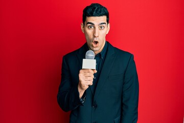 Handsome hispanic man holding reporter microphone scared and amazed with open mouth for surprise,...