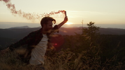 Fototapeta premium Male hipster running in field with smoke bomb. Smiling guy waving smoke grenade