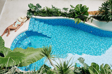 Beautiful young woman sunbathing with hat on her face by swimming pool of spa hotel, view from above