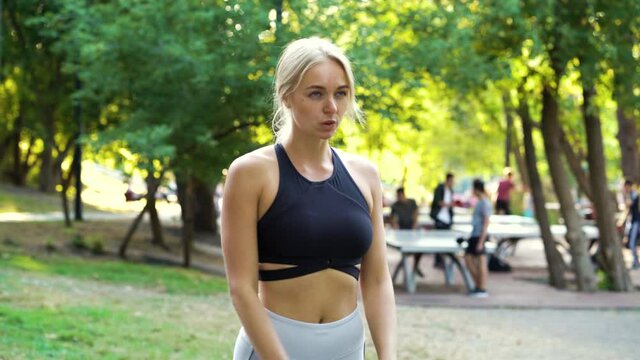 Blonde woman wearing black top and white leggings standing in park and looking concentrated, blurred people on background playing ping pong. Female jogger getting ready to start. Concept of sport
