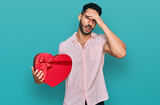 Hispanic man with beard holding valentine gift stressed and frustrated with hand on head, surprised and angry face