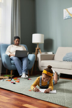 Vertical portrait of cute African-American girl drawing while lying on floor in home interior with mother working with laptop in background, copy space