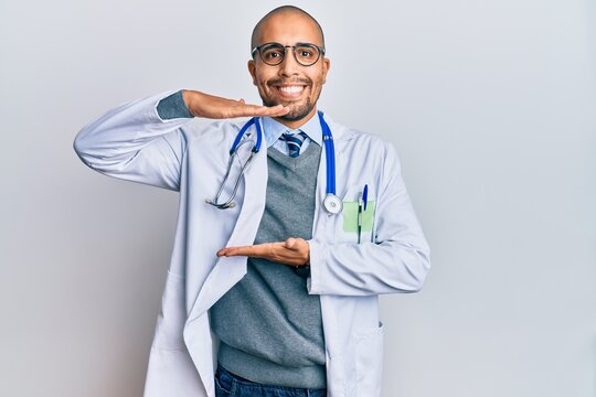Hispanic adult man wearing doctor uniform and stethoscope gesturing with hands showing big and large size sign, measure symbol. smiling looking at the camera. measuring concept.