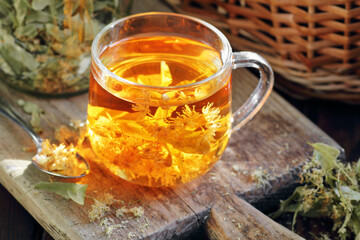 Linden herbal tea with flowers in a glass cup on wooden rustic board with lime blossom dry herb nearby, closeup, drink for relaxation, mental health,  naturopathy, homeopathy and folk medicine concept