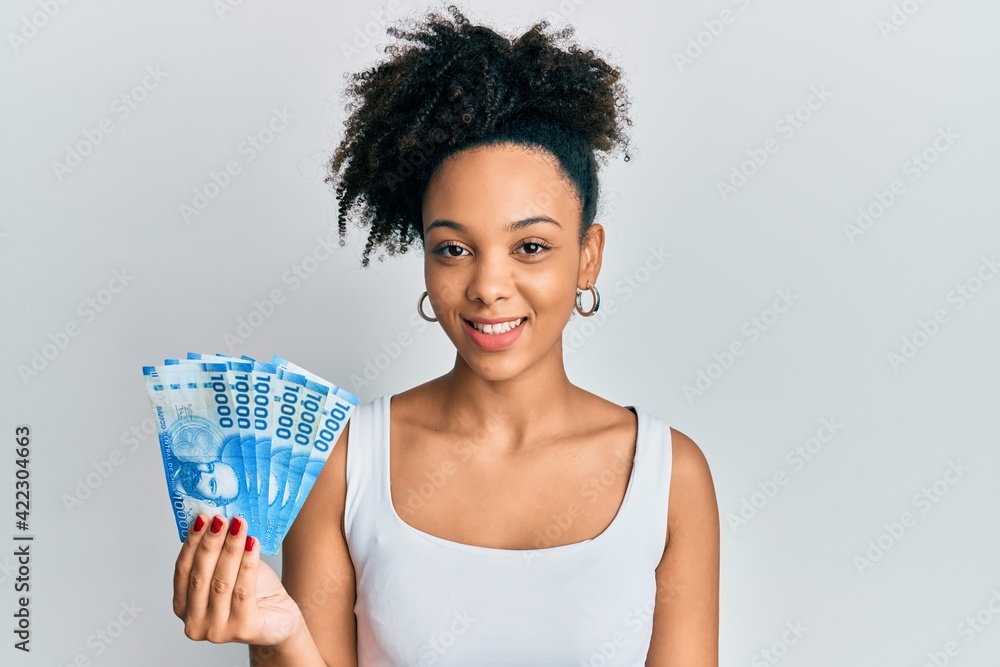 Poster Young african american girl holding 10000 chilean pesos looking positive and happy standing and smiling with a confident smile showing teeth