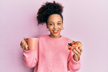 Young african american girl drinking a cup of coffee and eating sweet winking looking at the camera...