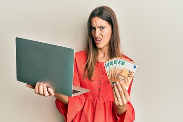 Young beautiful woman holding laptop and 50 euros in shock face, looking skeptical and sarcastic, surprised with open mouth