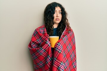 Young brunette woman with curly hair wrapped in a red warm red blanket drinking coffee clueless and...