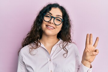 Young brunette woman with curly hair wearing casual clothes and glasses showing and pointing up with fingers number three while smiling confident and happy.