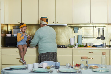 Full length portrait of cute African-American girl sitting on kitchen counter while helping...