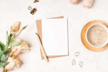 Craft paper envelope with white blank paper note mockup with pencil, flowers, cup of coffee, tag, twine on white background. Flat lay, top view. Invitation, package and letter.