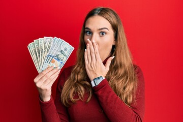 Young blonde woman holding dollars covering mouth with hand, shocked and afraid for mistake....