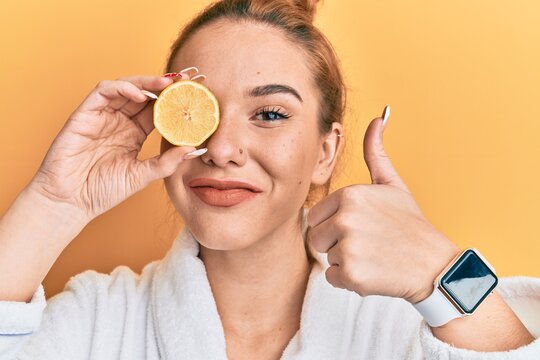 Young blonde woman wearing bathrobe holding lemon over eye smiling happy and positive, thumb up doing excellent and approval sign