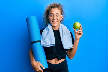 Beautiful caucasian teenager girl holding yoga mat and green apple winking looking at the camera...