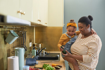 Waist up portrait of happy African-American mother holding daughter while cooking together in...