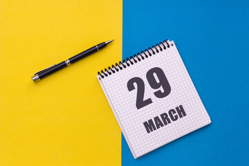 March 29th. Day 29 of month, calendar date. A notebook with a spiral and a pen lies on a yellow-blue background, flat lay, copy space
