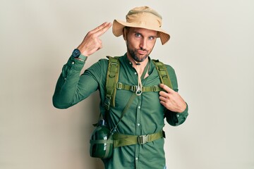 Fototapeta premium Handsome man with beard wearing explorer hat and backpack shooting and killing oneself pointing hand and fingers to head like gun, suicide gesture.