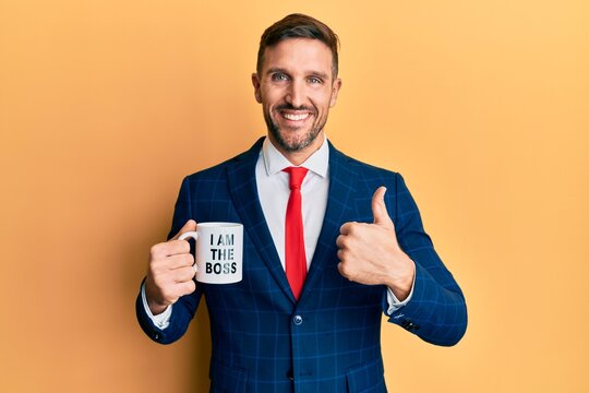 Handsome business man with beard drinking from i am the boss coffee cup smiling happy and positive, thumb up doing excellent and approval sign