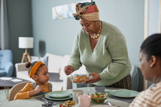 Portrait of African-American family enjoying dinner together in cozy home interior, focus on grandmother serving food to cute little girl