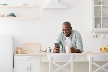 Happy african american man using smartphone and having morning coffee in the kitchen