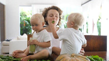 Caucasian baby sitting on the kitchen table among green leaves drinking green smoothie they've just...