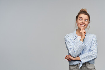 Portrait of attractive, dreaming girl with blond hair bun. Wearing blue striped knotted shirt. Touching her chin and watching curious to the left at copy space, isolated over grey background